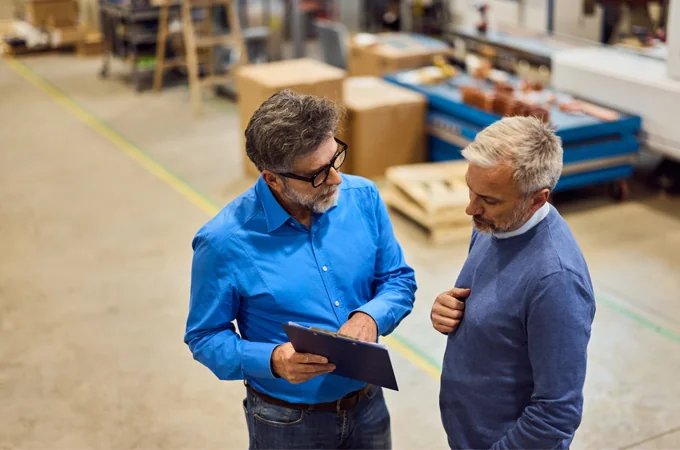 An advisor reviews a clipboard with a colleague on a manufacturing shop floor.