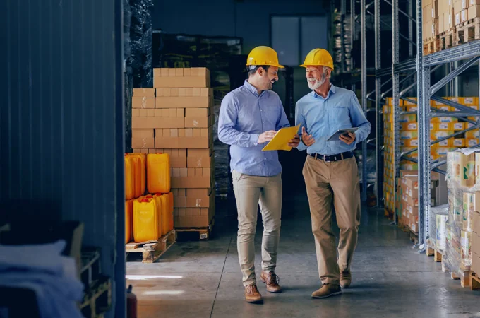 Two workers in hard hats walk a warehouse aisle reviewing notes on a clipboard and tablet.