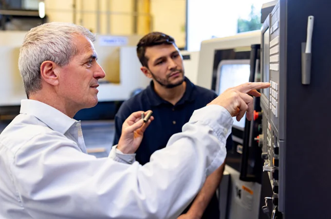 An instructor points out a control on a CNC machine panel while a technician looks on.