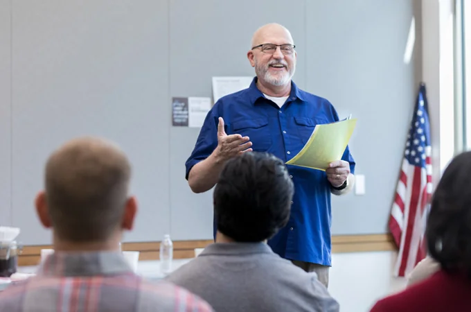An instructor holding handouts speaks to a seated classroom of trainees.